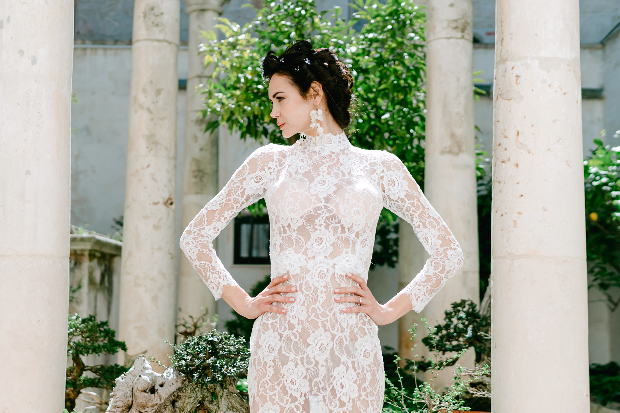 A woman wearing a long-sleeved, white lace dress stands confidently with hands on her hips, posing outdoors in front of stone columns and greenery.