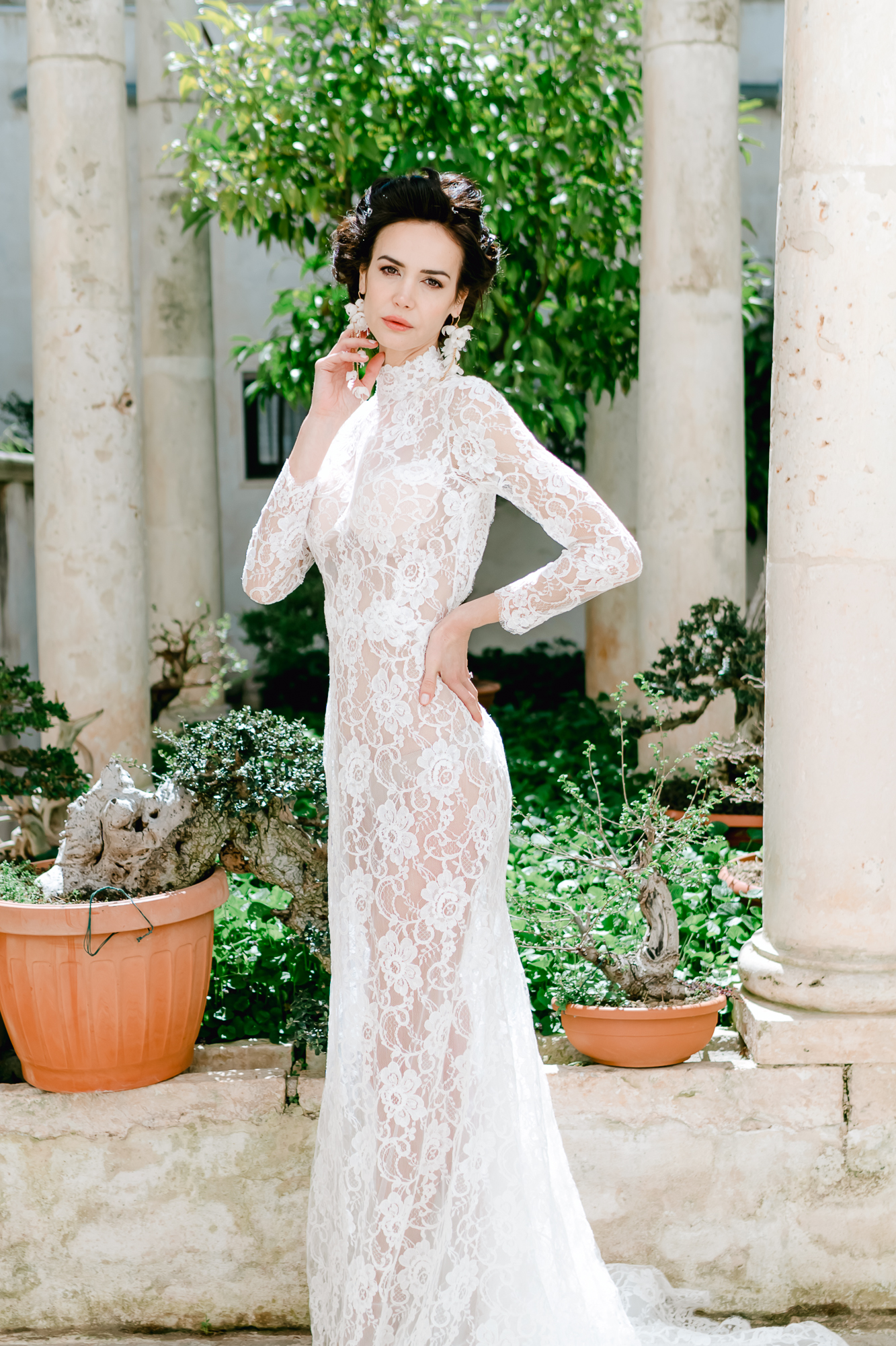 A woman in a long-sleeved, sheer lace wedding dress stands in a sunlit courtyard with stone columns and potted plants, posing with one hand on her hip and the other lightly touching her face.