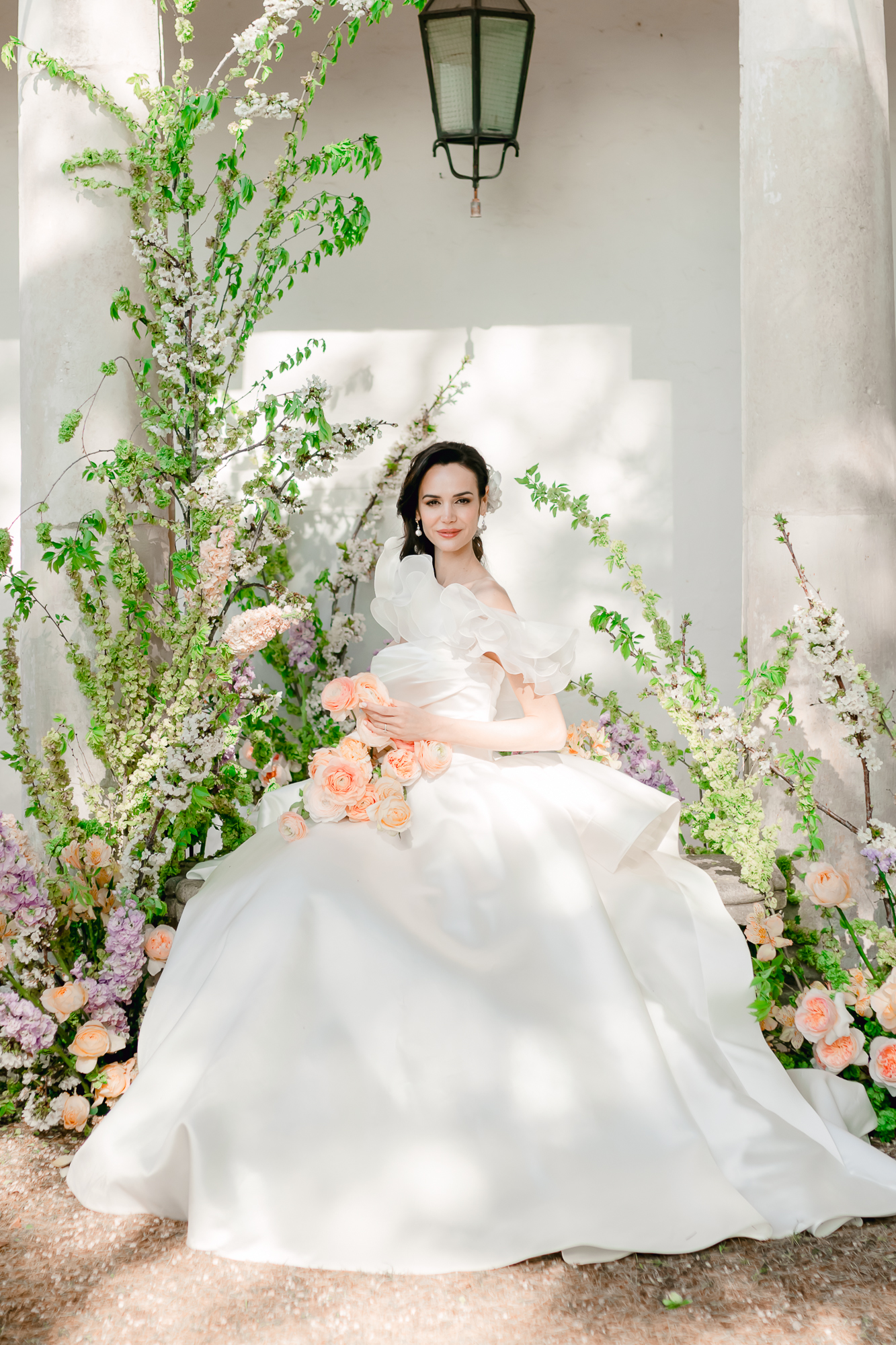 A bride in a white wedding gown sits outdoors, surrounded by lush greenery and pastel flowers, with sunlight casting soft shadows on the wall behind her. She holds a bouquet of peach and pink roses.