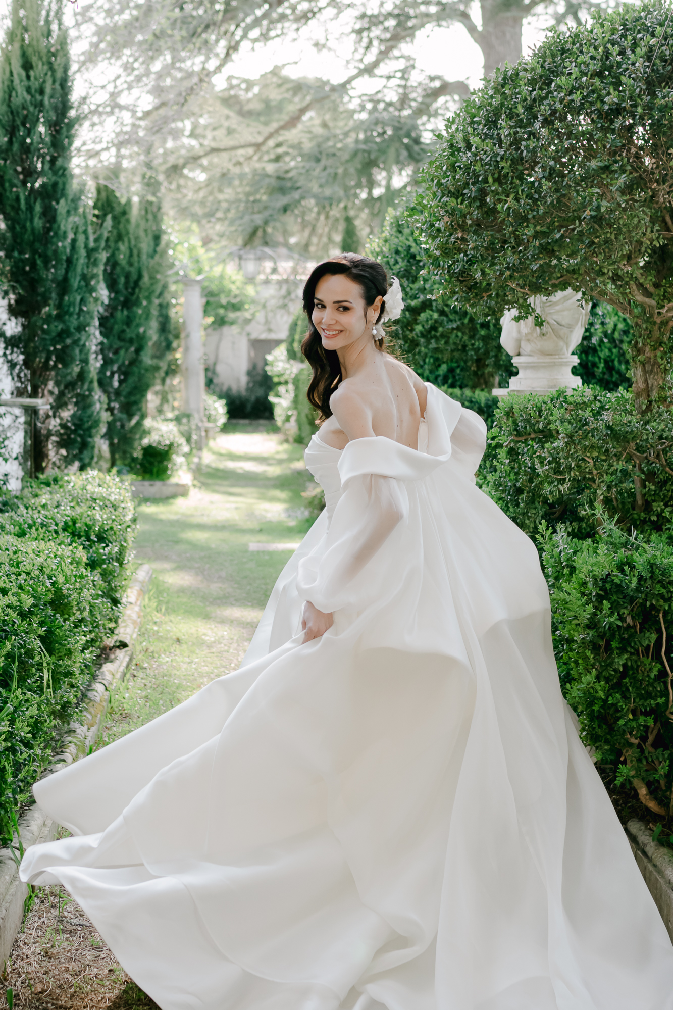 A woman in a flowing white wedding dress smiles as she looks over her shoulder, walking along a garden path lined with green hedges and trees.