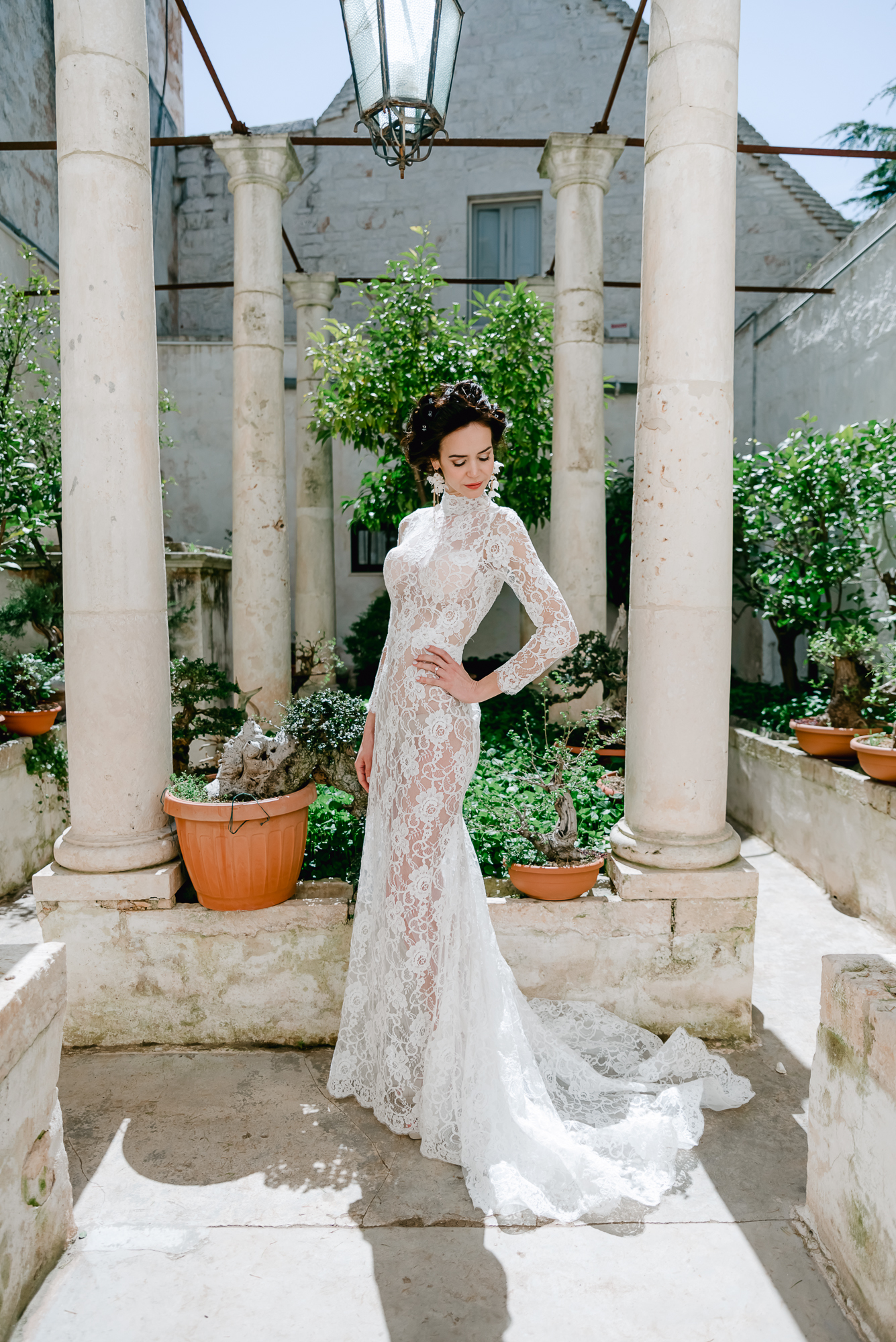 A woman in a long, white lace wedding dress stands confidently among stone columns in a sunlit garden courtyard, surrounded by potted plants and greenery.