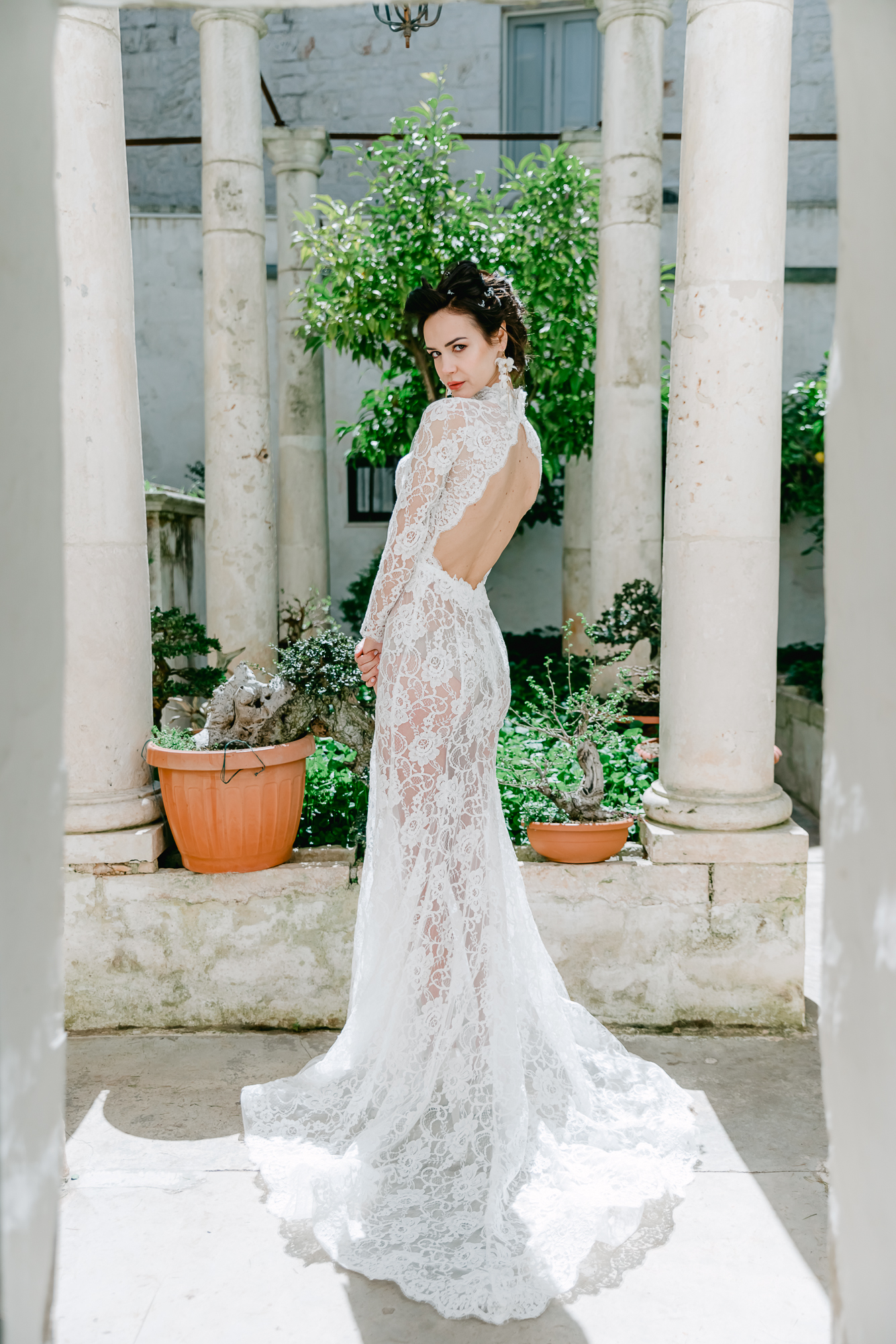 A woman in a long, lace, backless white bridal gown stands between stone columns in a sunlit courtyard, looking over her shoulder with greenery and potted plants in the background.