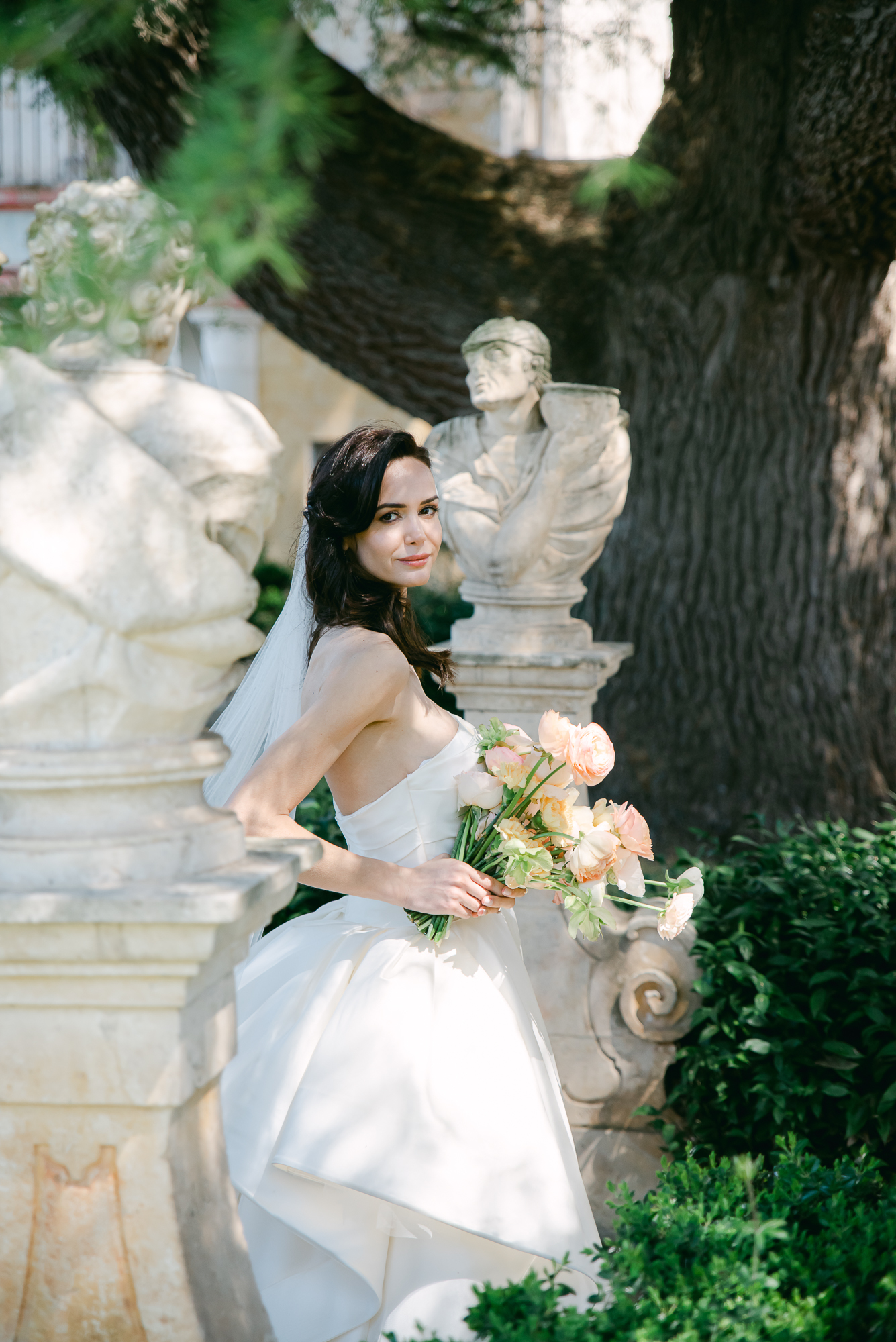 A bride in a white dress and veil holds a bouquet of peach roses, standing outdoors between two stone bust sculptures with greenery and a large tree in the background.