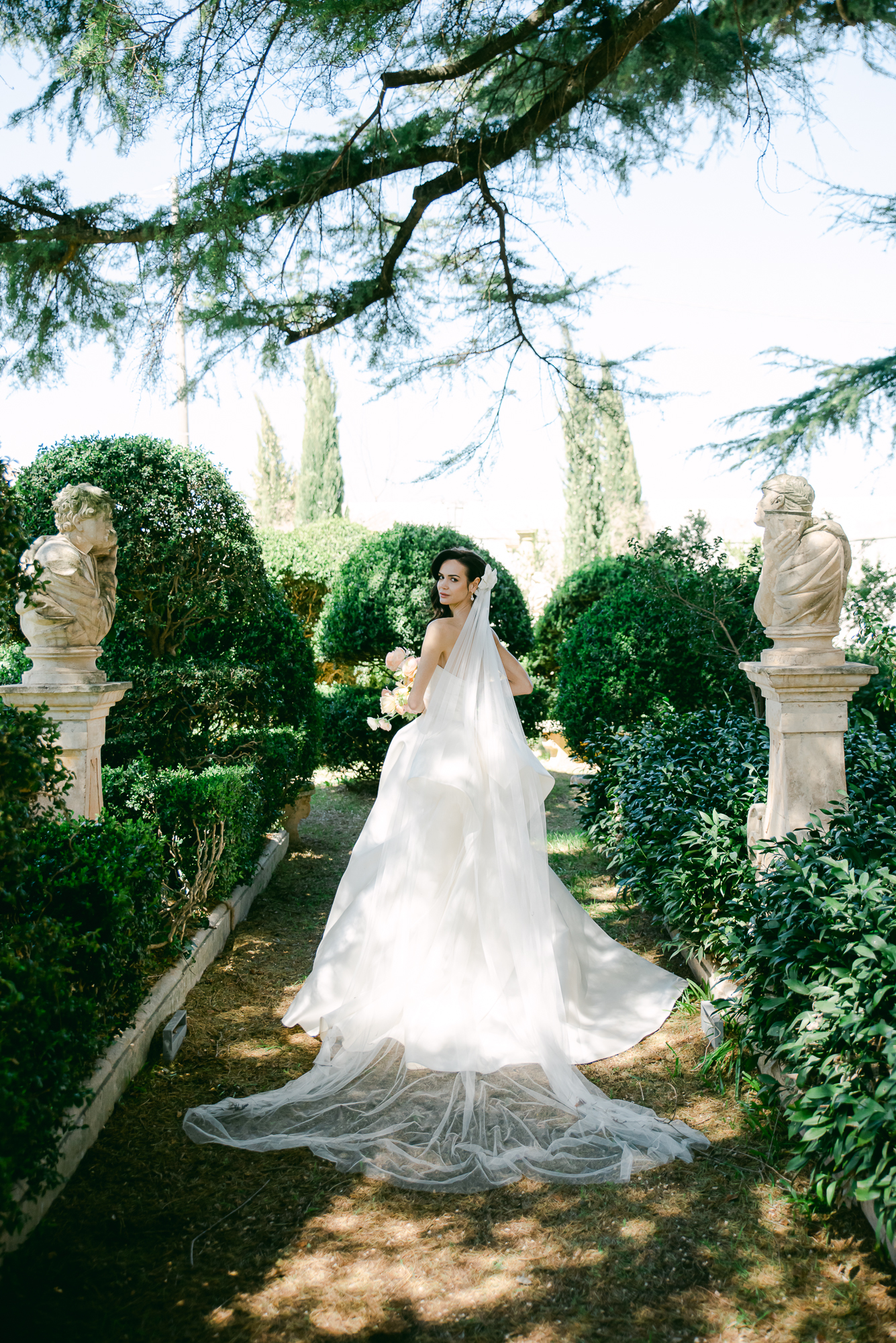 A bride in a flowing white gown stands on a garden path bordered by green bushes and stone statues, with sunlight filtering through tree branches above.