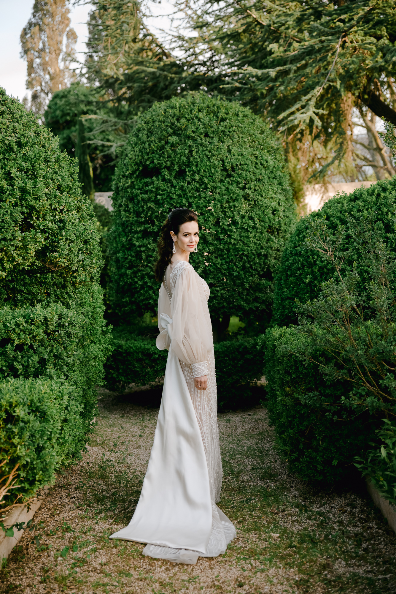 A woman in a long white gown stands on a gravel path between tall, manicured green bushes, looking over her shoulder in a lush garden setting.