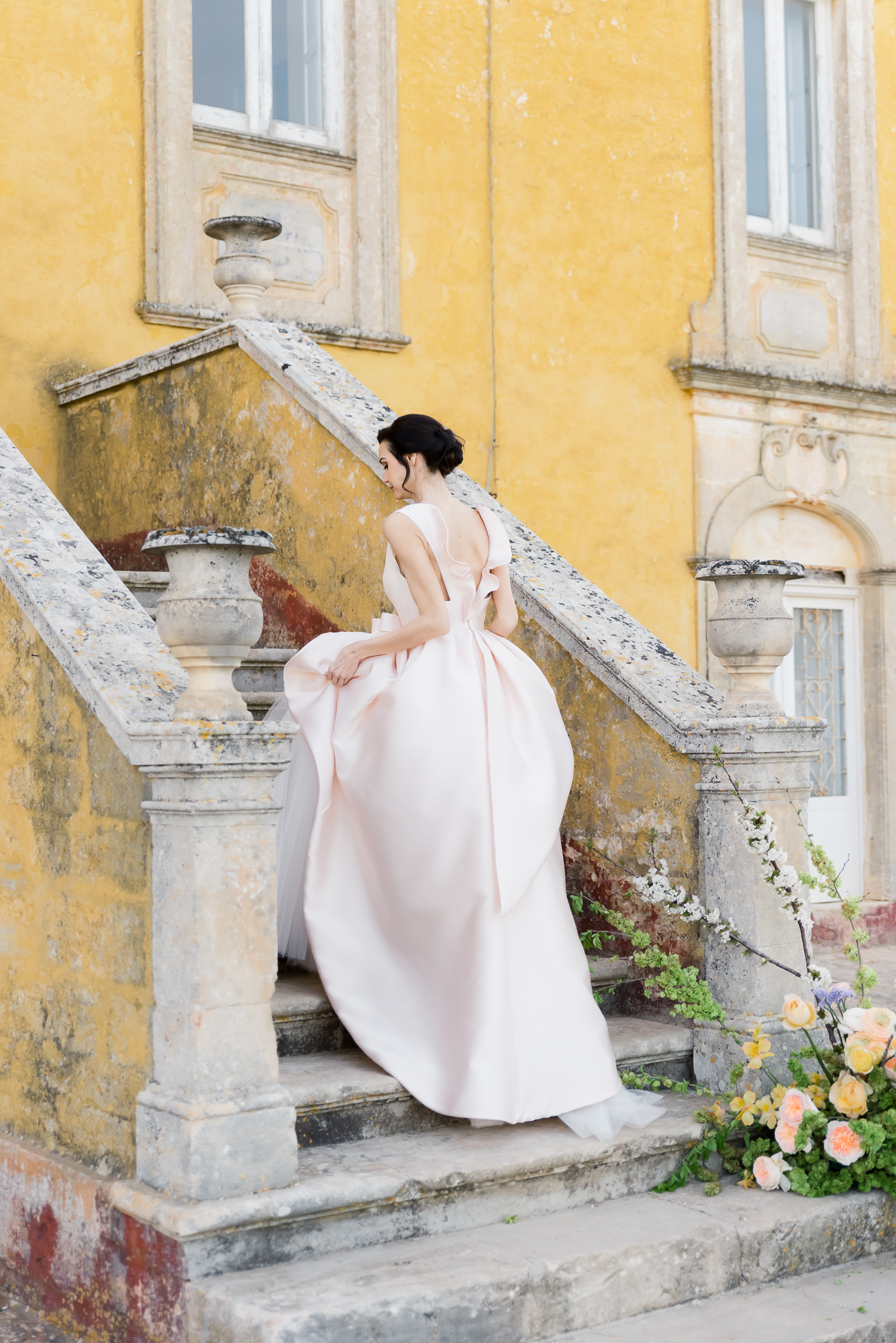 A woman in a pale pink, elegant gown walks up stone stairs outside a yellow historic building, holding her dress to avoid the flowers and greenery arranged on the steps.