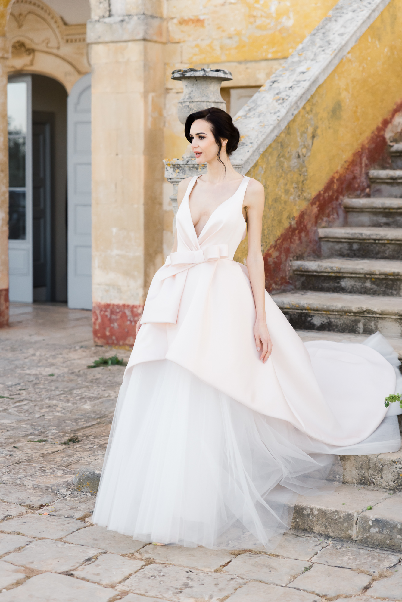 A woman in a white, sleeveless, layered ball gown with a deep V-neckline stands next to stone stairs outside a weathered, historic building with a pale yellow wall and arched doorway.