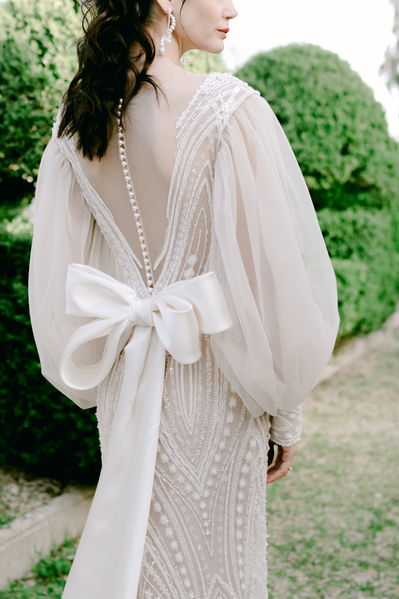 A woman in a beaded, sheer-sleeved wedding dress with a deep V-back, pearl buttons, and a large white bow stands outside near green bushes.
