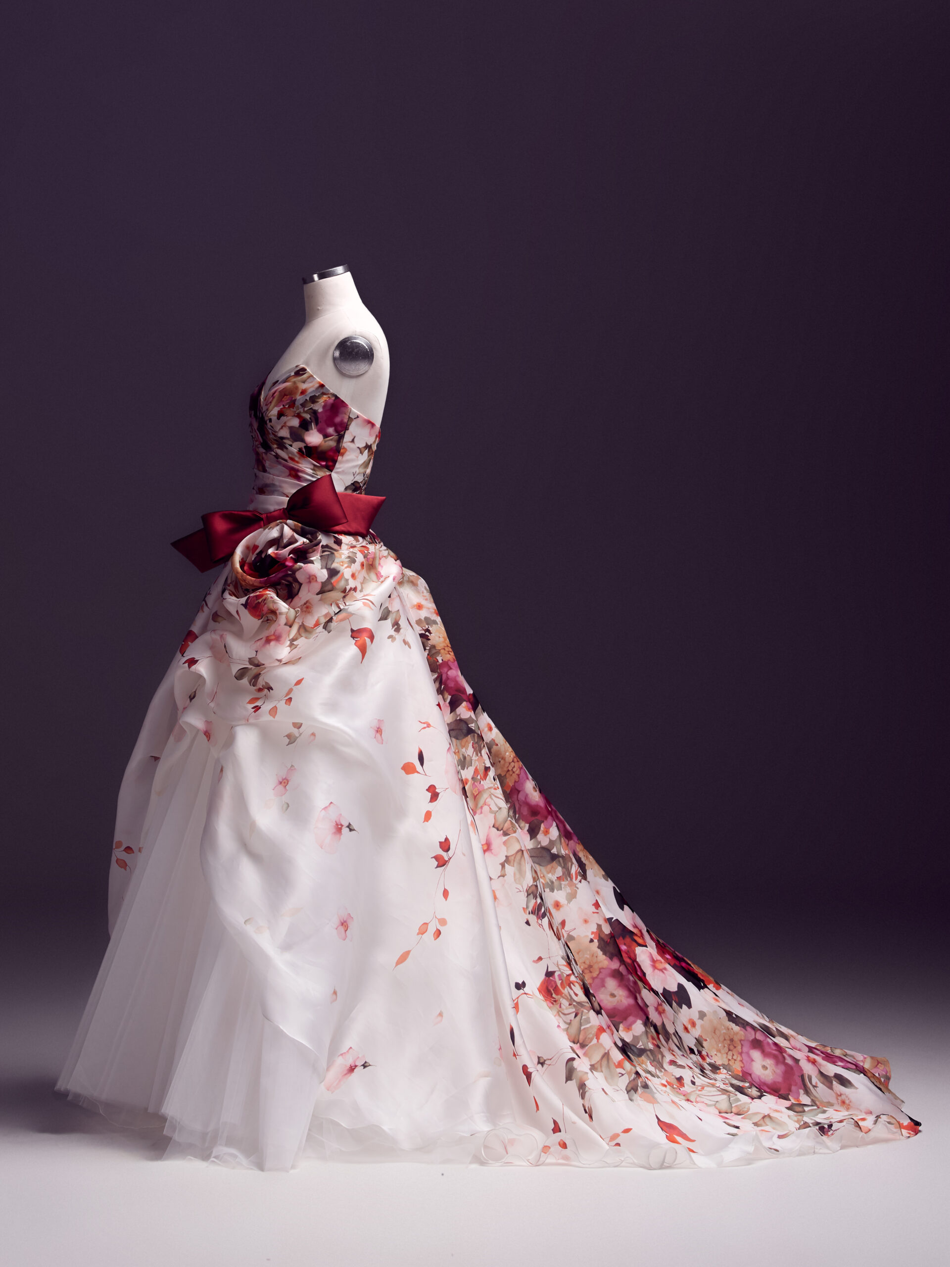 A white ball gown on a dress form features a floral print, a strapless bodice, a dramatic red bow at the waist, and a flowing skirt with layers and a floral train, set against a dark background.