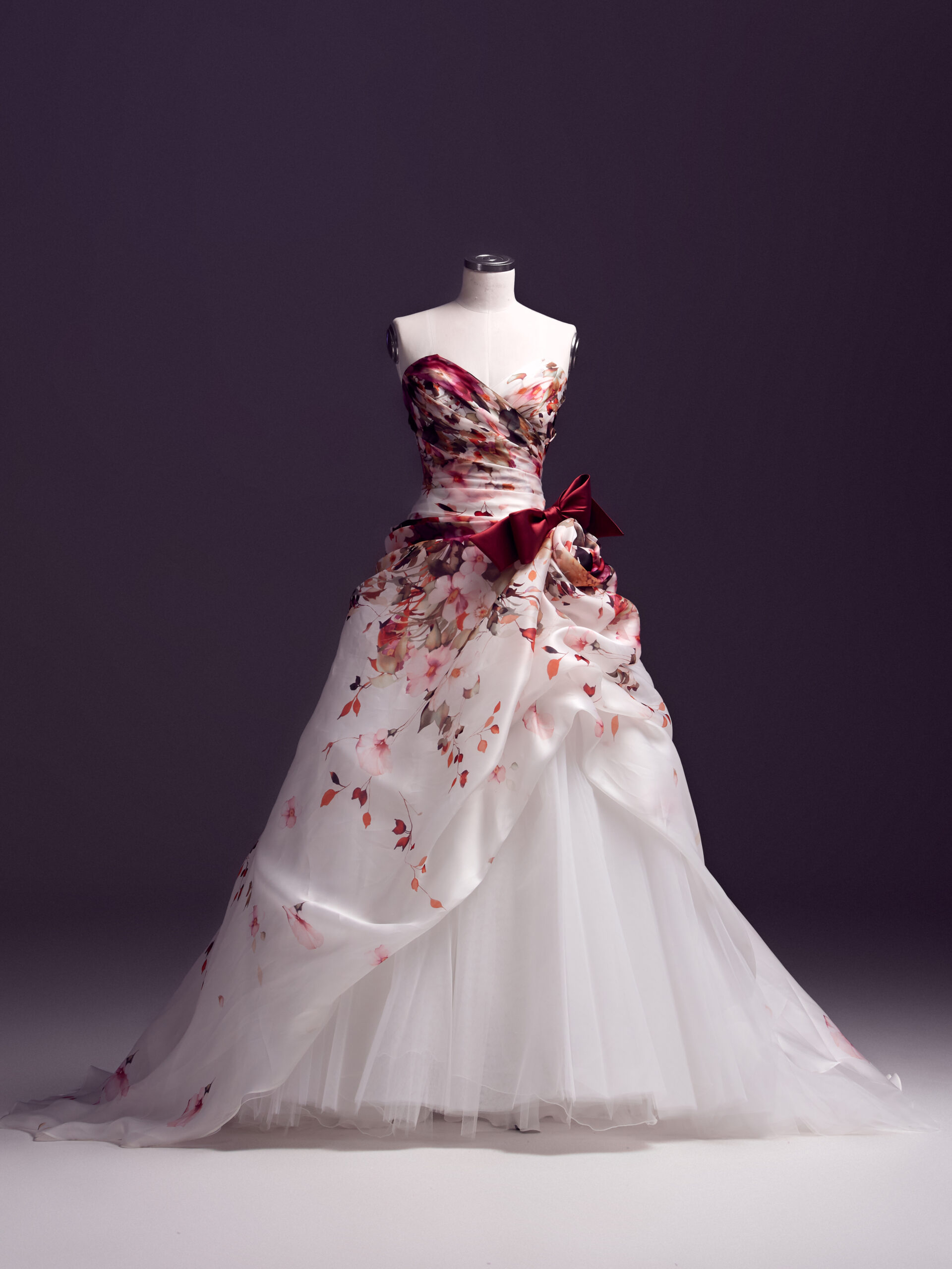 A strapless ball gown with a sweetheart neckline, featuring floral patterns in shades of pink and red, a burgundy ribbon at the waist, and a flowing layered white skirt displayed on a dress form against a dark background.