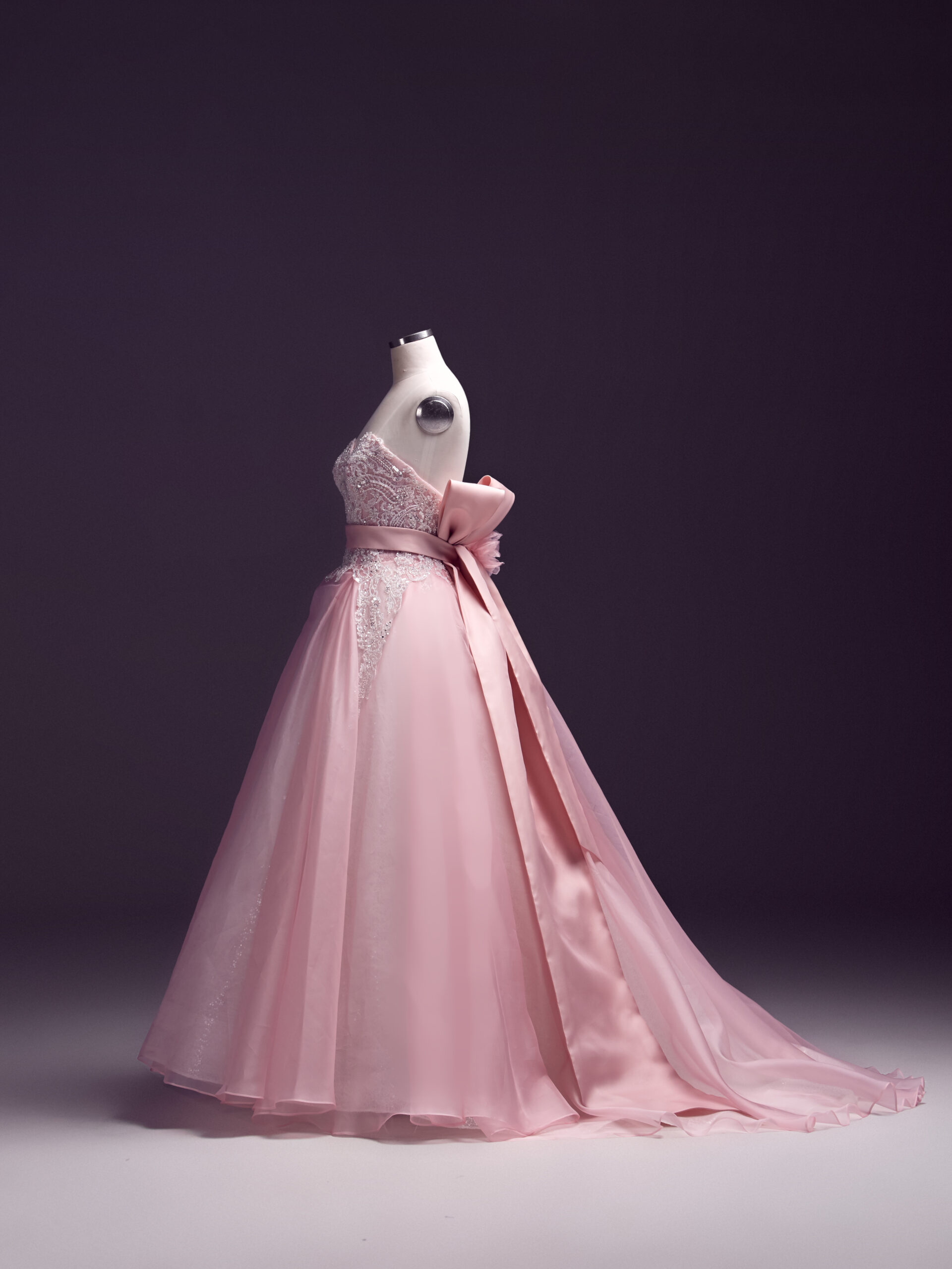 A strapless pink ball gown with a fitted bodice, lace details, and a large decorative bow at the waist, displayed on a dress form against a dark background. The dress features a long, flowing skirt and train.