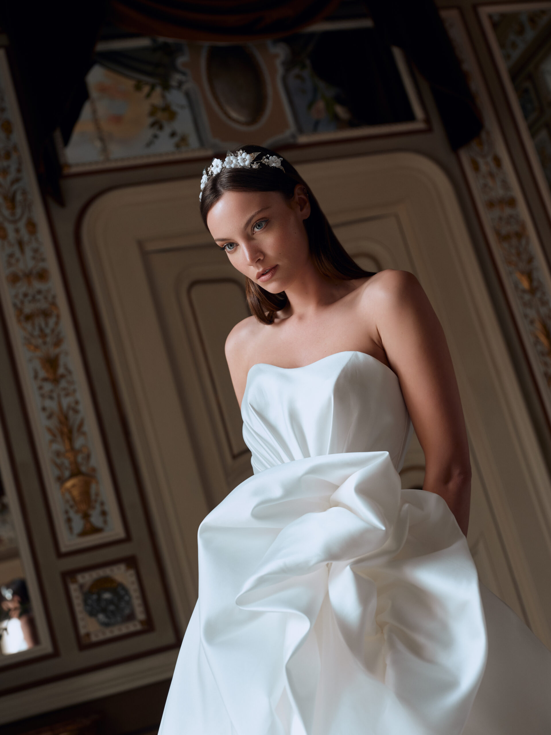 A woman in a strapless white gown with a large, sculpted detail at the waist stands indoors, wearing a jeweled headband. She is in an ornately decorated room with detailed molding and elegant wall designs.