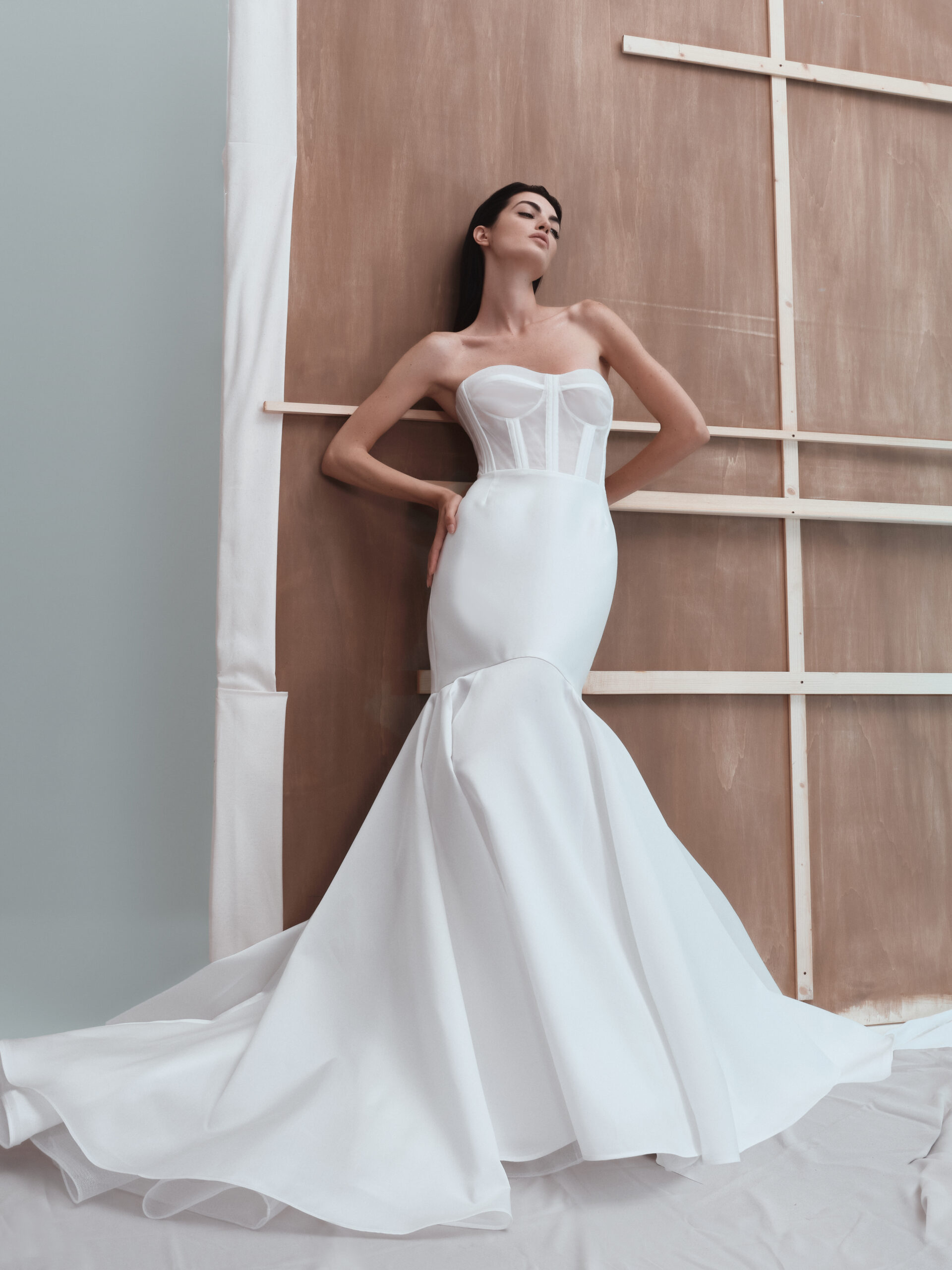 A woman poses in a strapless white mermaid wedding dress with a structured corset bodice and dramatic flared skirt, standing against a wooden backdrop with white fabric partially draped to the side.