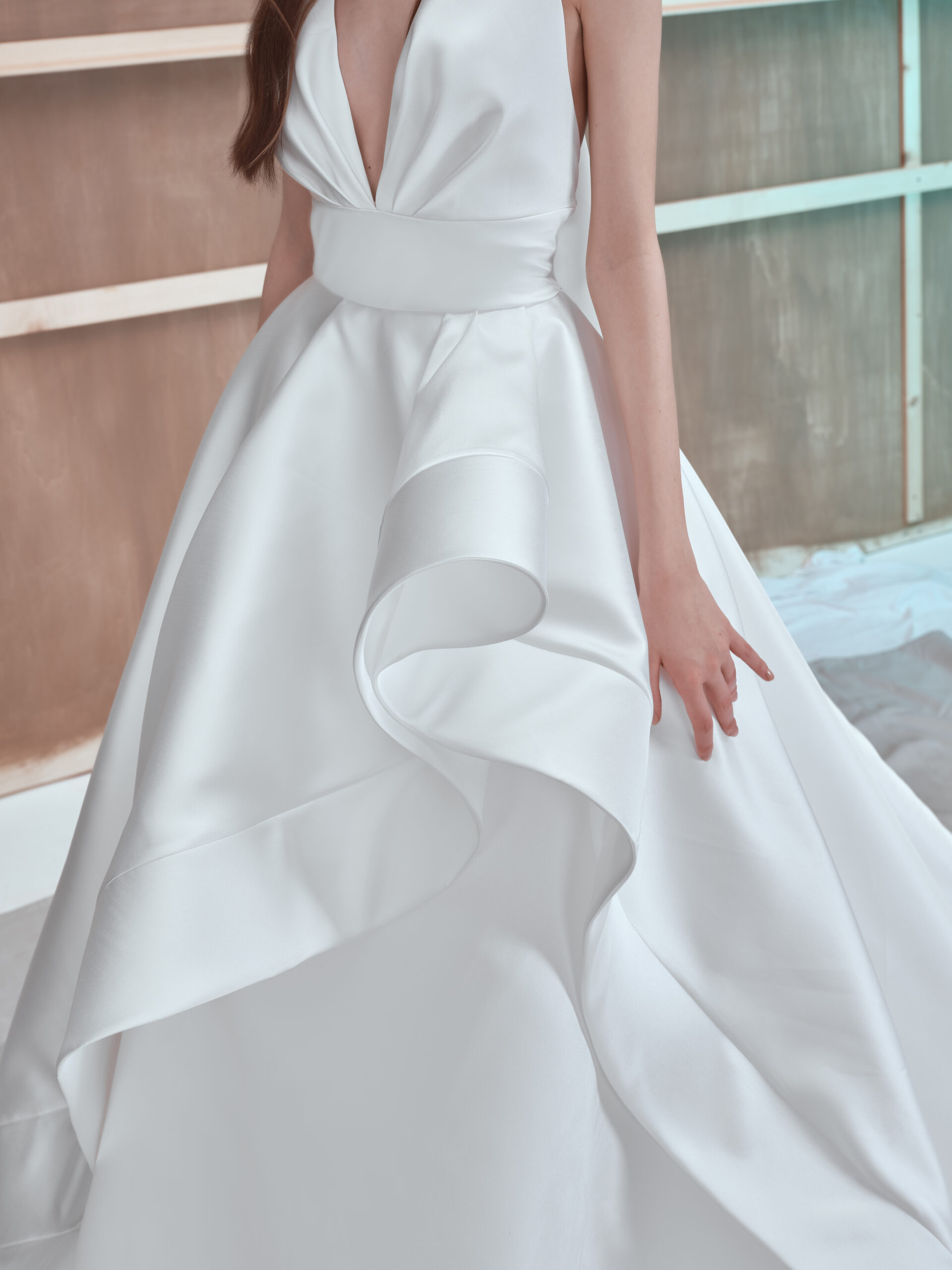 A woman wearing a sleeveless white satin gown with a deep V-neck and dramatic, sculpted ruffle detail on the skirt. Her face is not visible; she stands against a neutral, modern background.