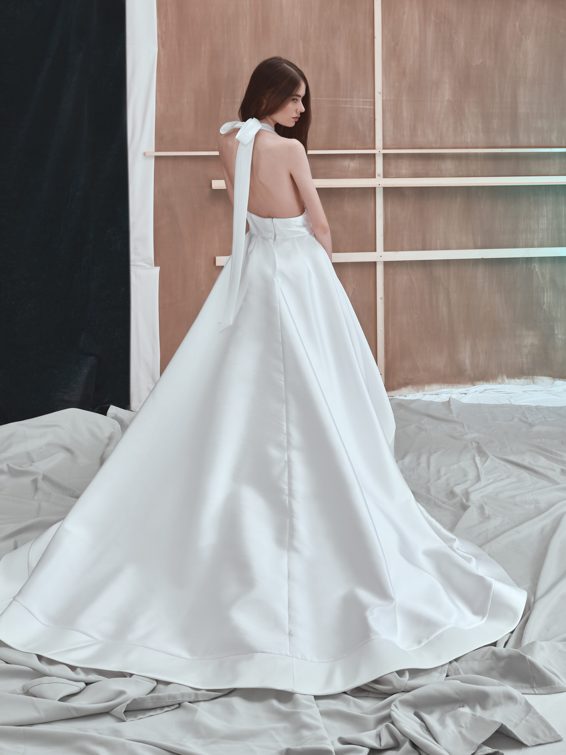 A woman stands on a draped fabric floor wearing a long, elegant white backless gown with a bow at the neck, facing away from the camera against a simple wooden and black backdrop.