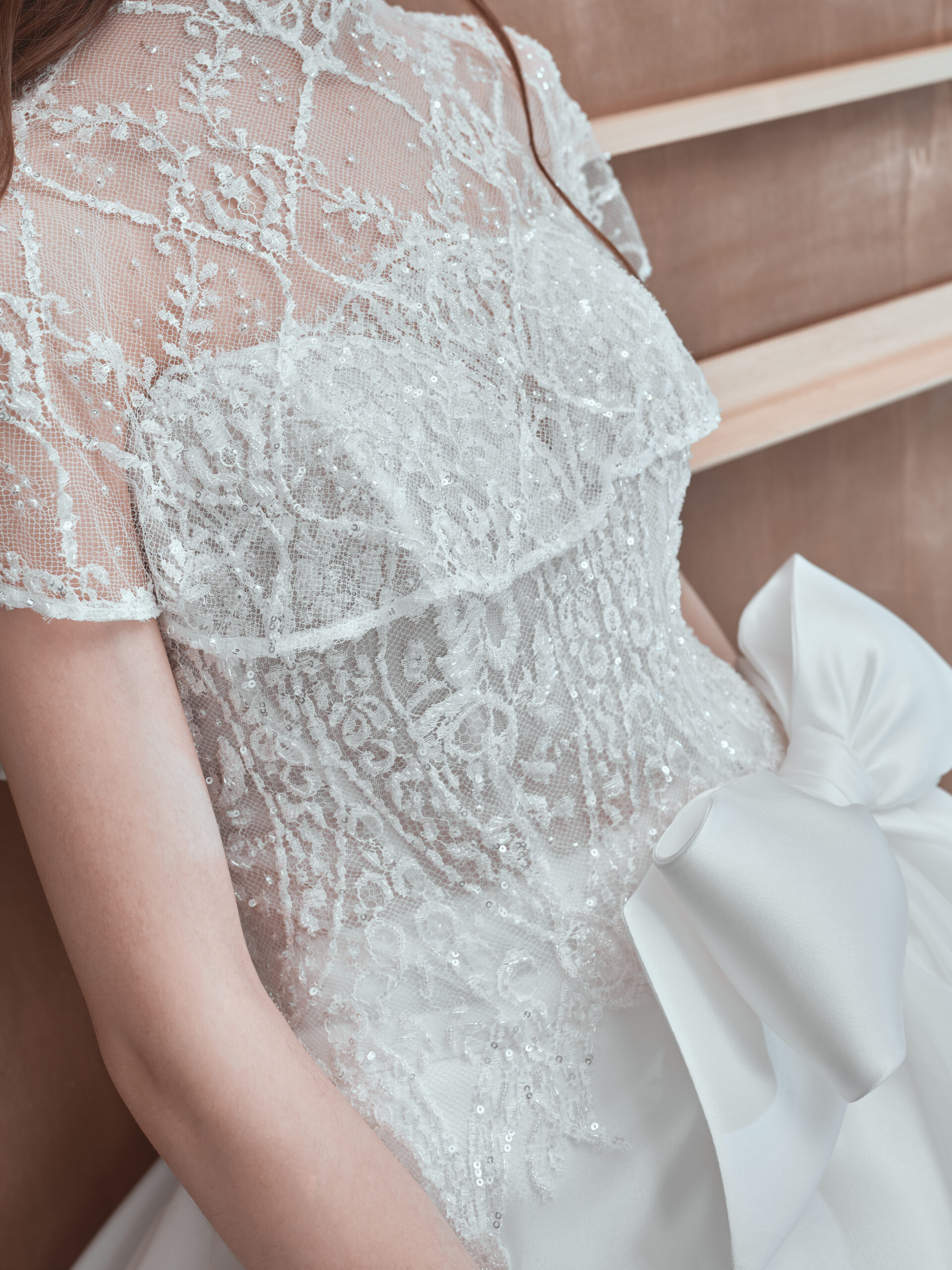 A close-up of a woman wearing a white lace wedding dress with intricate embroidery and a large satin bow at the waist, standing near wooden shelves.