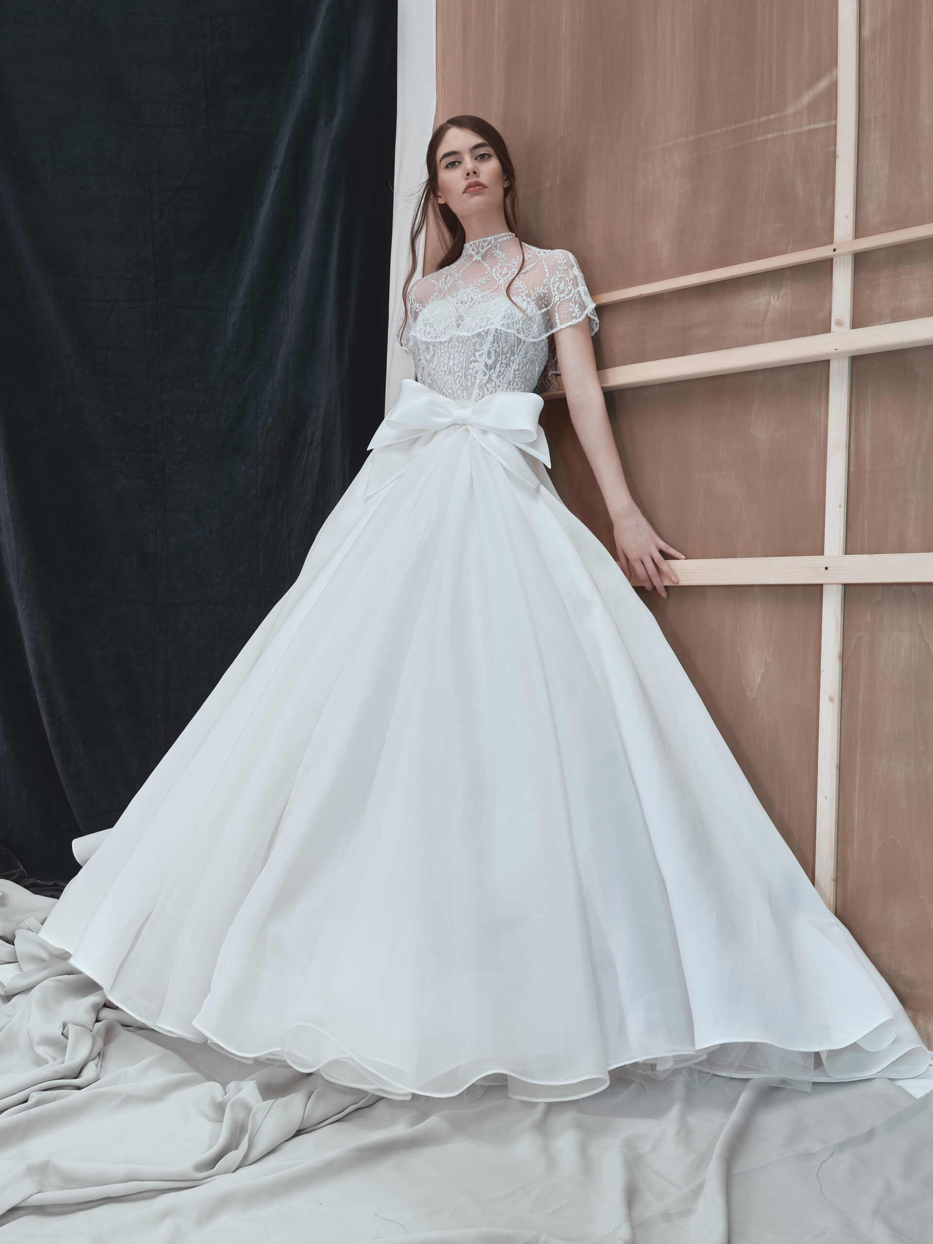 A woman in a white lace and satin ball gown with a large bow at the waist stands against a wooden and black draped backdrop, posing with her arms slightly extended and gazing ahead.