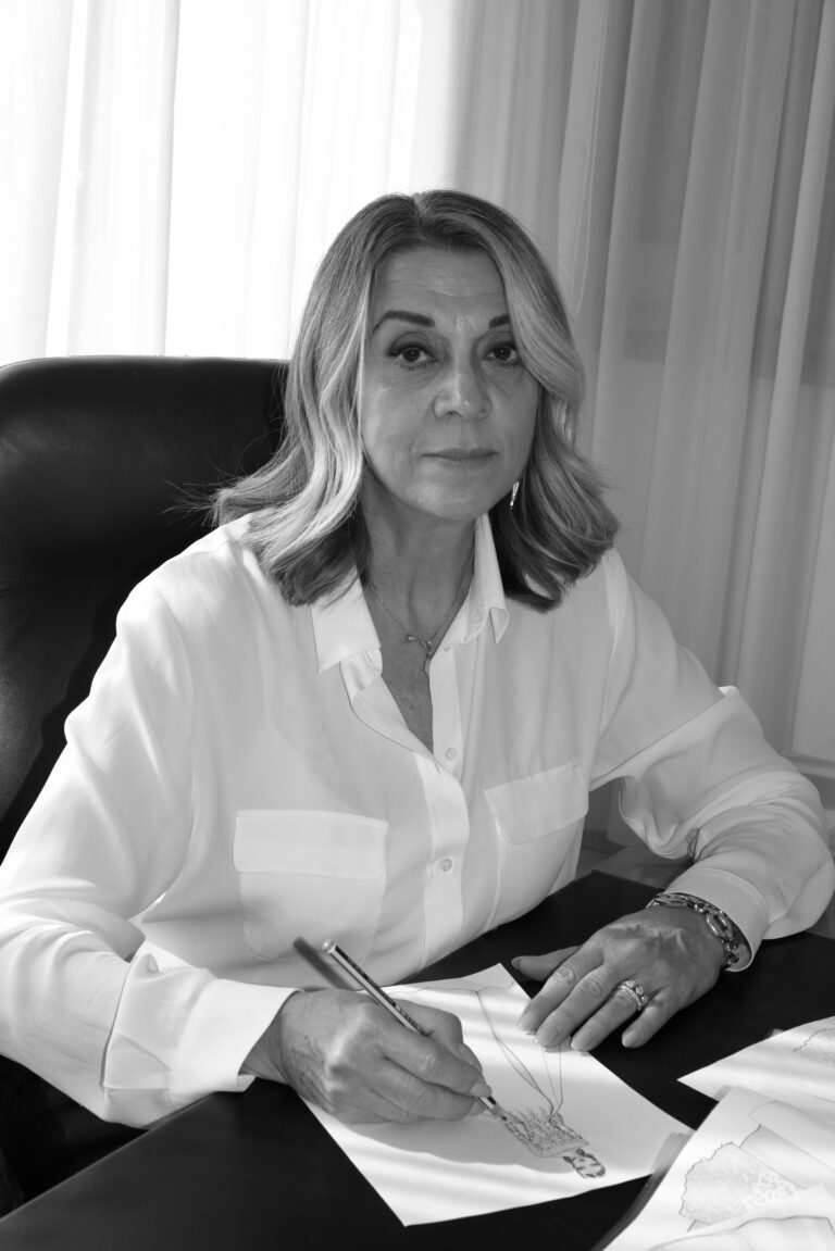 A woman with shoulder-length hair wearing a light blouse sits at a desk, looking at the camera while holding a pen and drawing on paper. The image is in black and white.