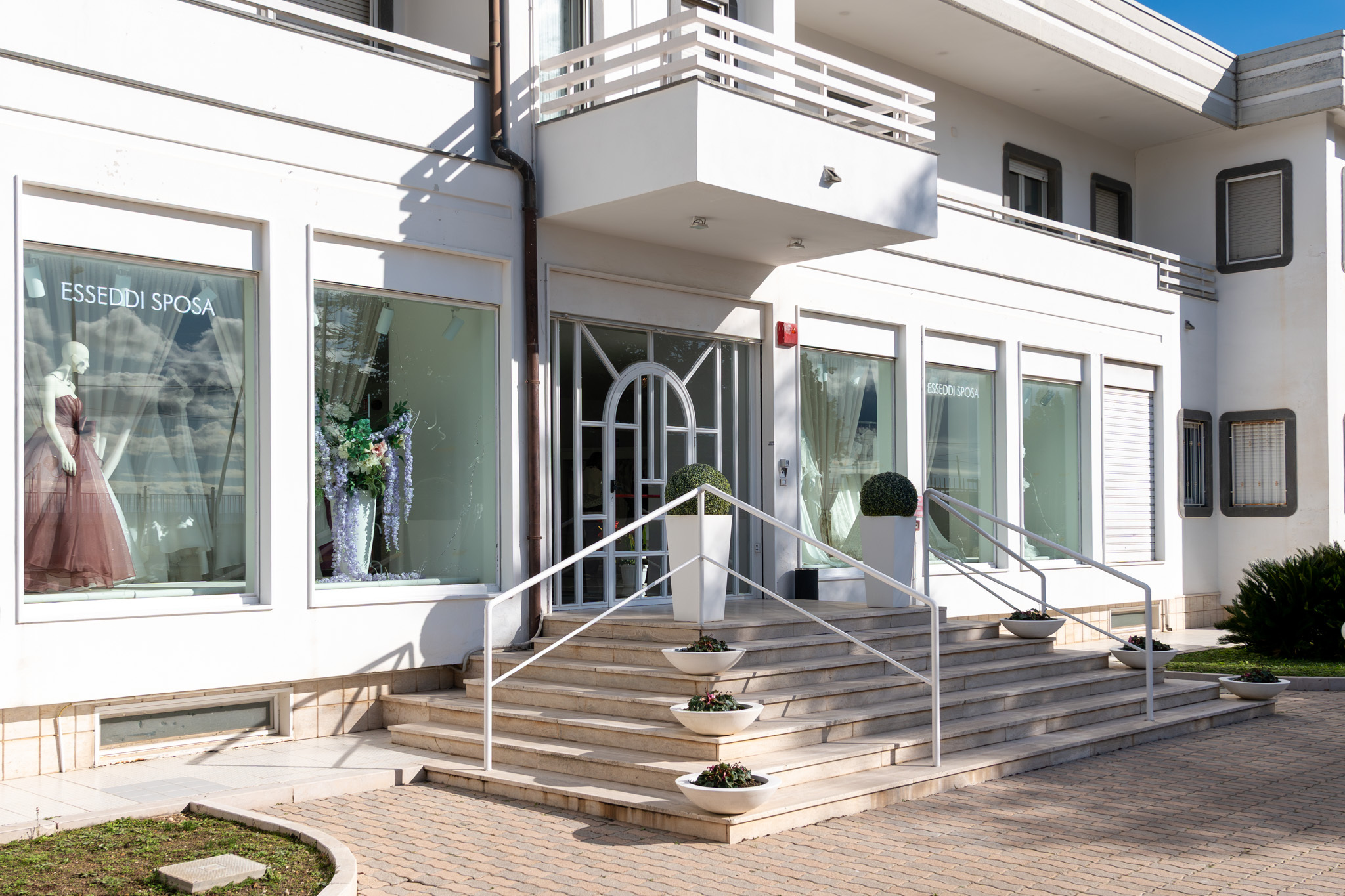 A modern bridal shop by Rosanna Perrone features large glass windows displaying mannequins in elegant dresses. Wide steps with potted plants lead to the entrance, and the building’s white exterior showcases clean lines.