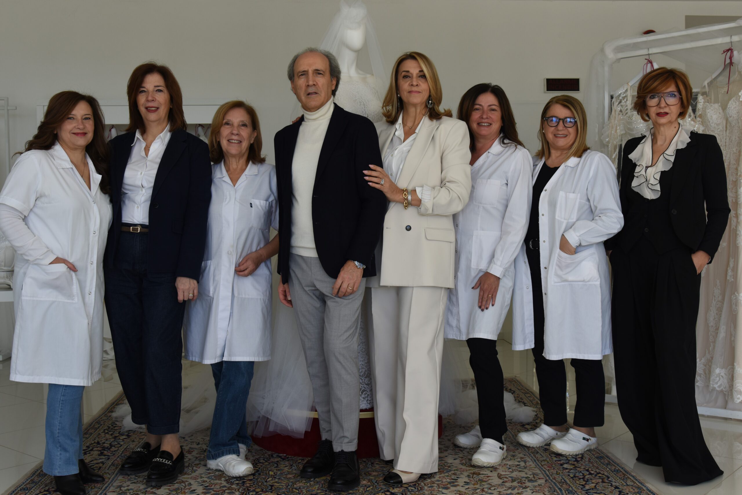 Seven women and one man stand together indoors, some wearing white lab coats and others in business attire, smiling at the camera. Dresses and fabric racks are visible in the background.